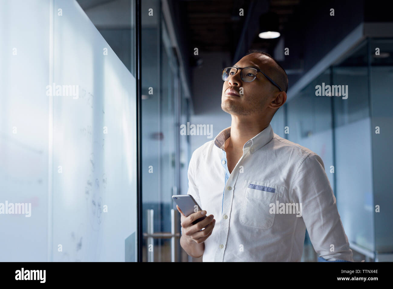 Thoughtful businessman holding smart phone en position debout par mur de verre at office Banque D'Images