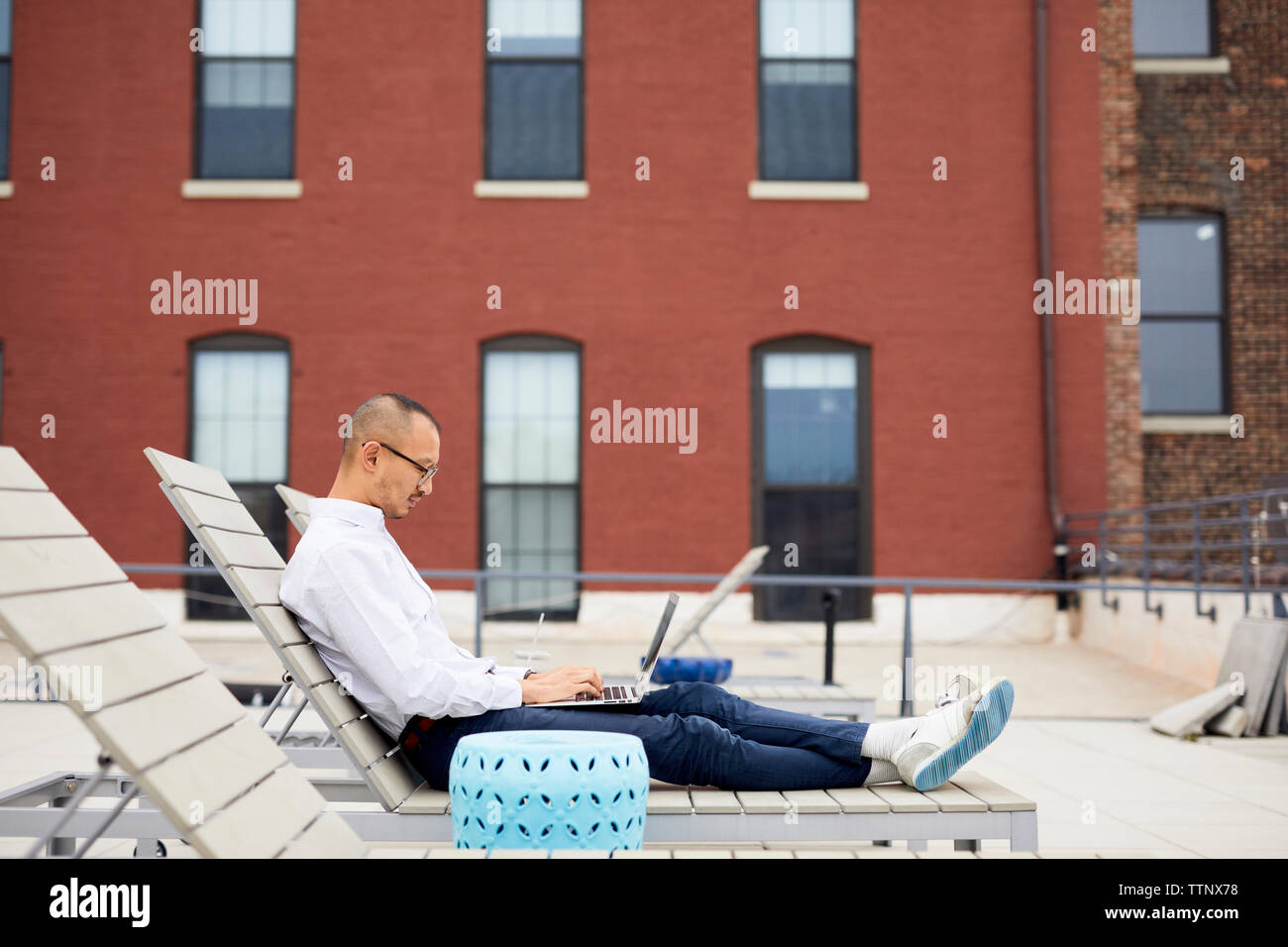 Vue latérale du businessman using laptop while sitting on lounge chair sur la terrasse de l'immeuble Banque D'Images