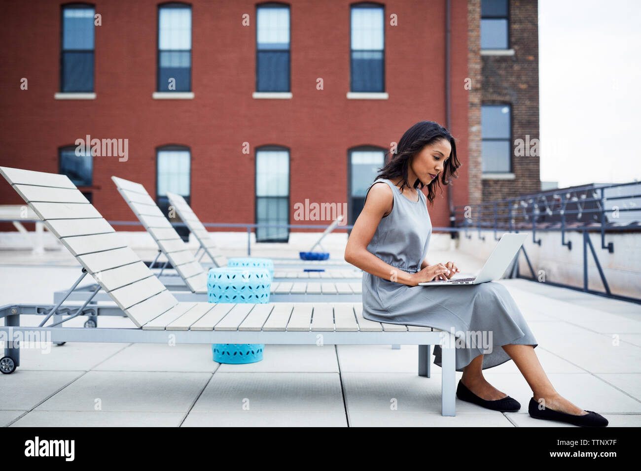 Toute la longueur de businesswoman using laptop while sitting on lounge chair à terrasse de l'immeuble Banque D'Images