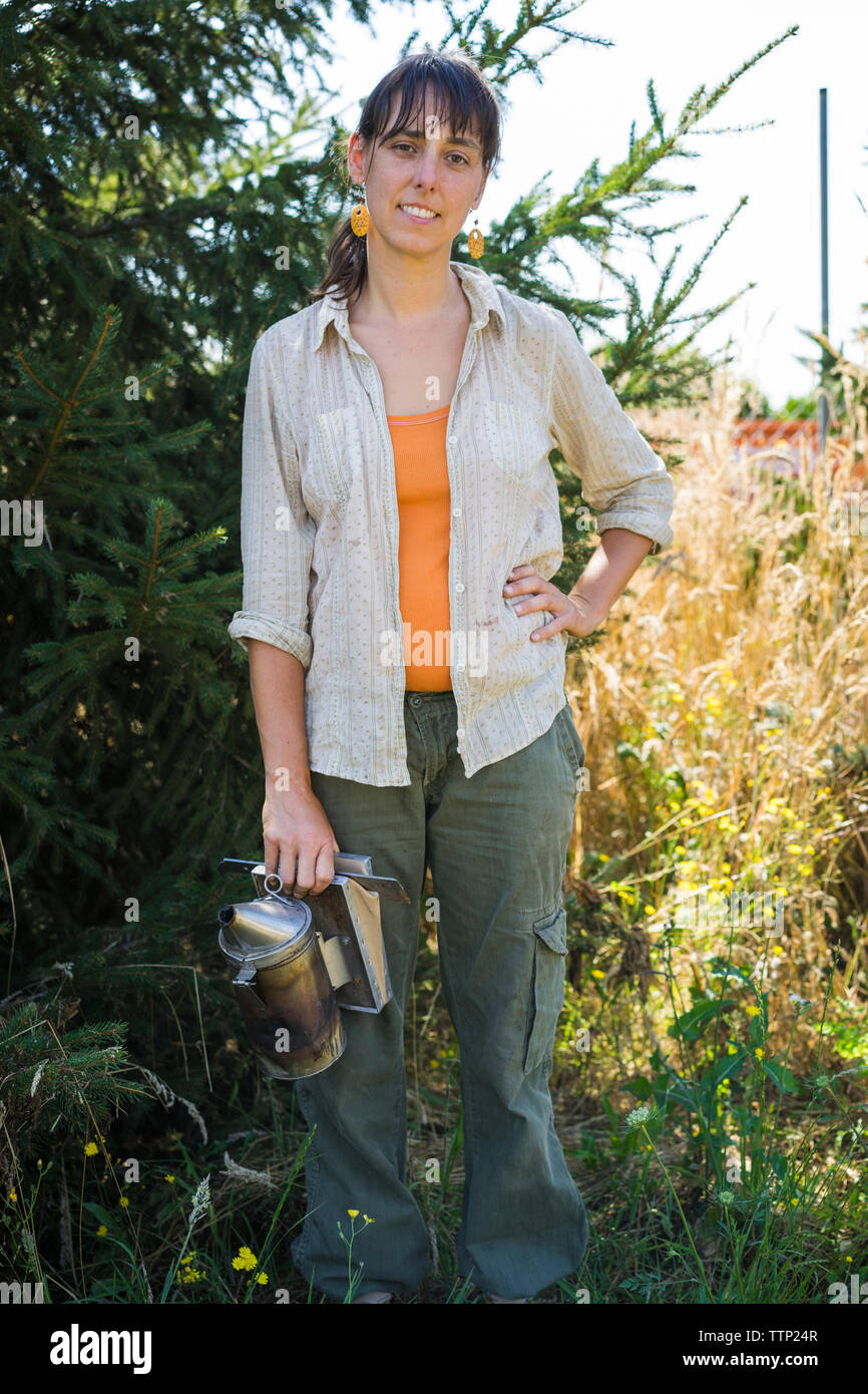 Portrait of smiling with hand on hip apiculteur abeille tenant debout fumeur contre des plantes sur le terrain Banque D'Images