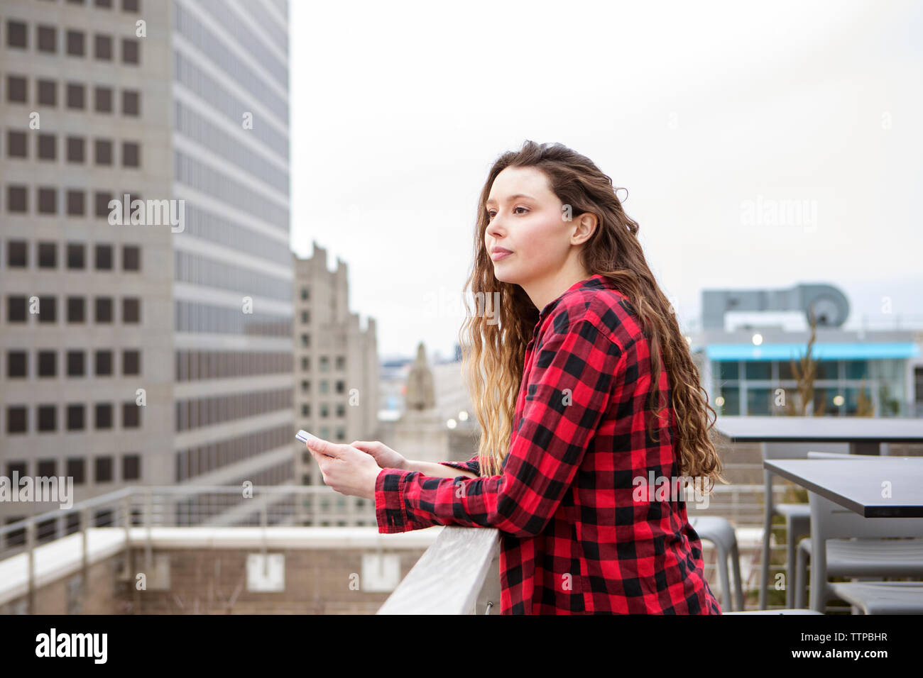 Young woman with smart phone standing by railing at Terrace cafe Banque D'Images