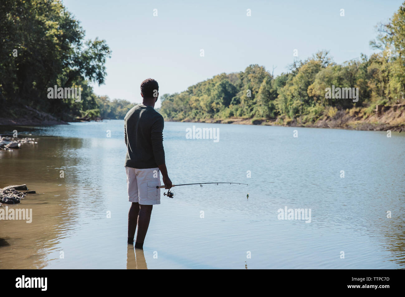 Vue arrière de l'homme tenant la canne à pêche en position debout dans le lac contre le ciel clair Banque D'Images
