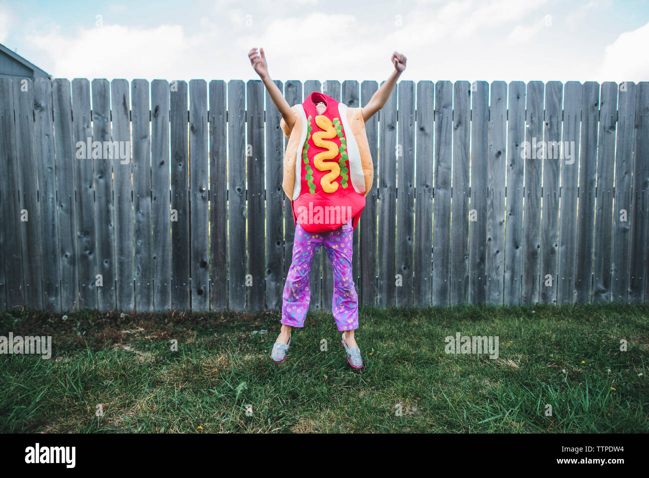 Fille aux bras levés de sauter tout en portant le costume de hot-dog contre clôture à yard Banque D'Images