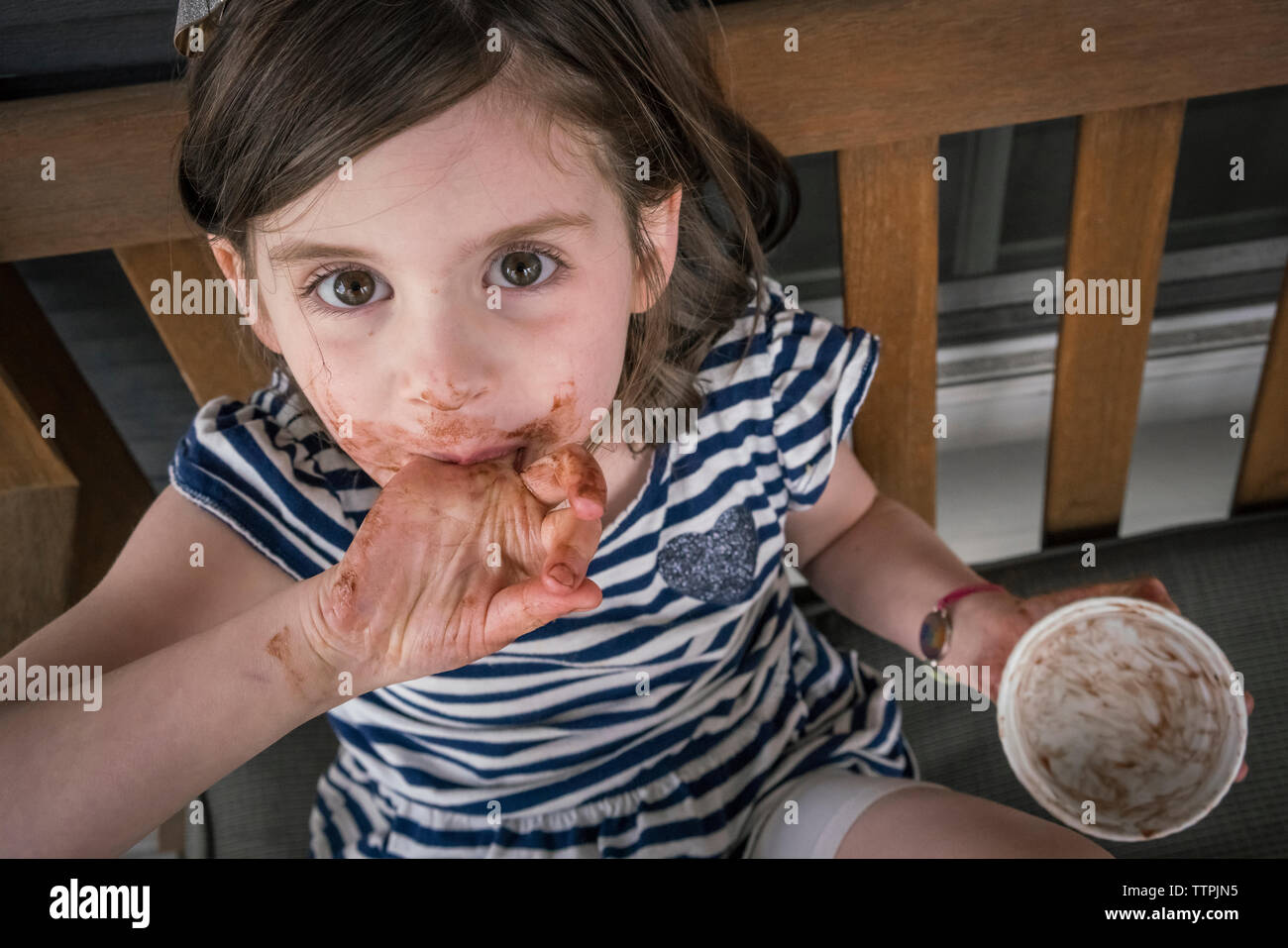 High angle Portrait of Girl with messy bouche manger du chocolat tout en restant assis sur une chaise à la maison Banque D'Images