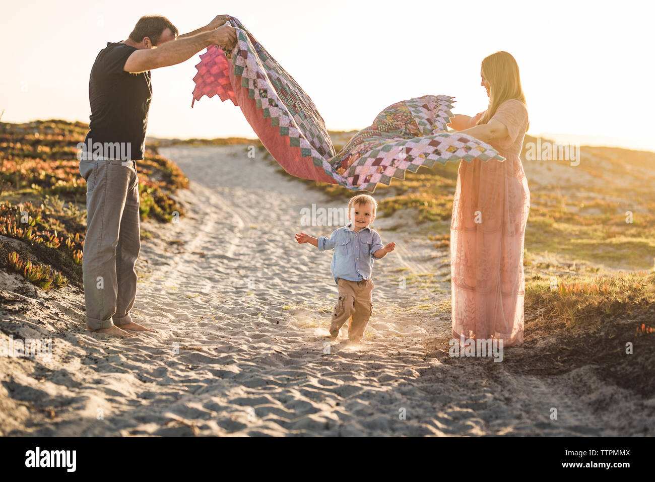 Vue latérale des parents holding couverture sur mignon fils d'exécution sur la plage de sable à contre ciel clair pendant le coucher du soleil Banque D'Images