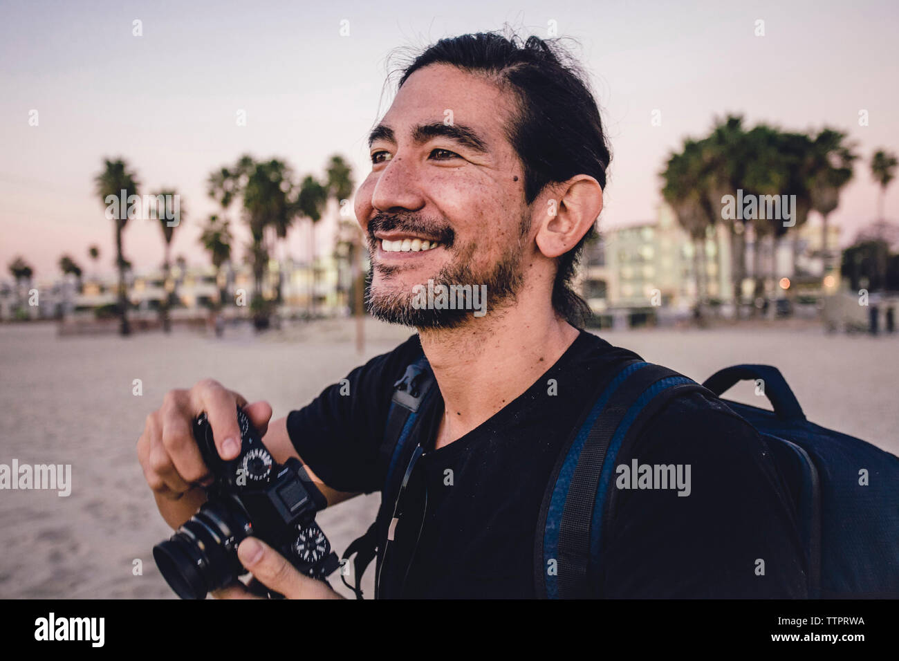 Close-up of smiling backpacker tenir l'appareil photo tout en regardant ailleurs at beach Banque D'Images