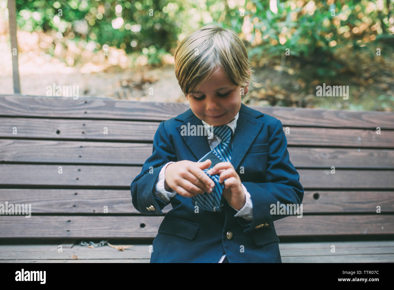 Cute boy in suit adjusting necktie while sitting on bench Banque D'Images