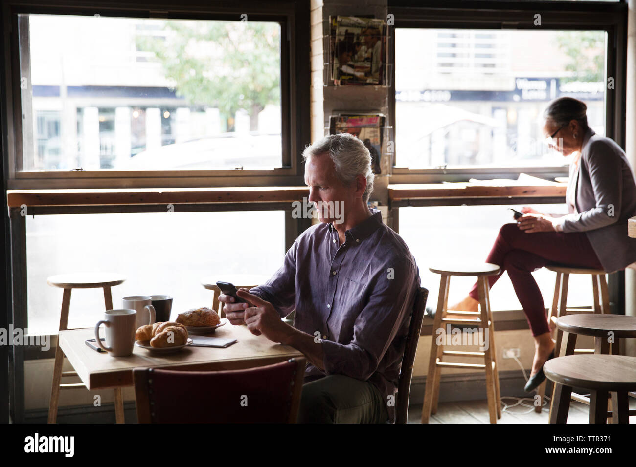 Les clients utilisant phone while sitting at table par windows dans cafe Banque D'Images