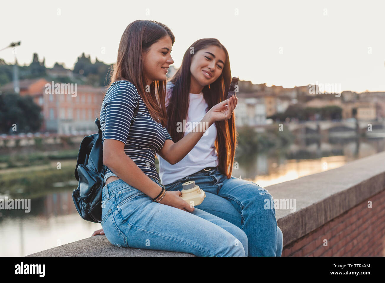 Smiling female friends looking at instant de transfert d'impression tout en restant assis sur mur de retenue par canal en ville Banque D'Images