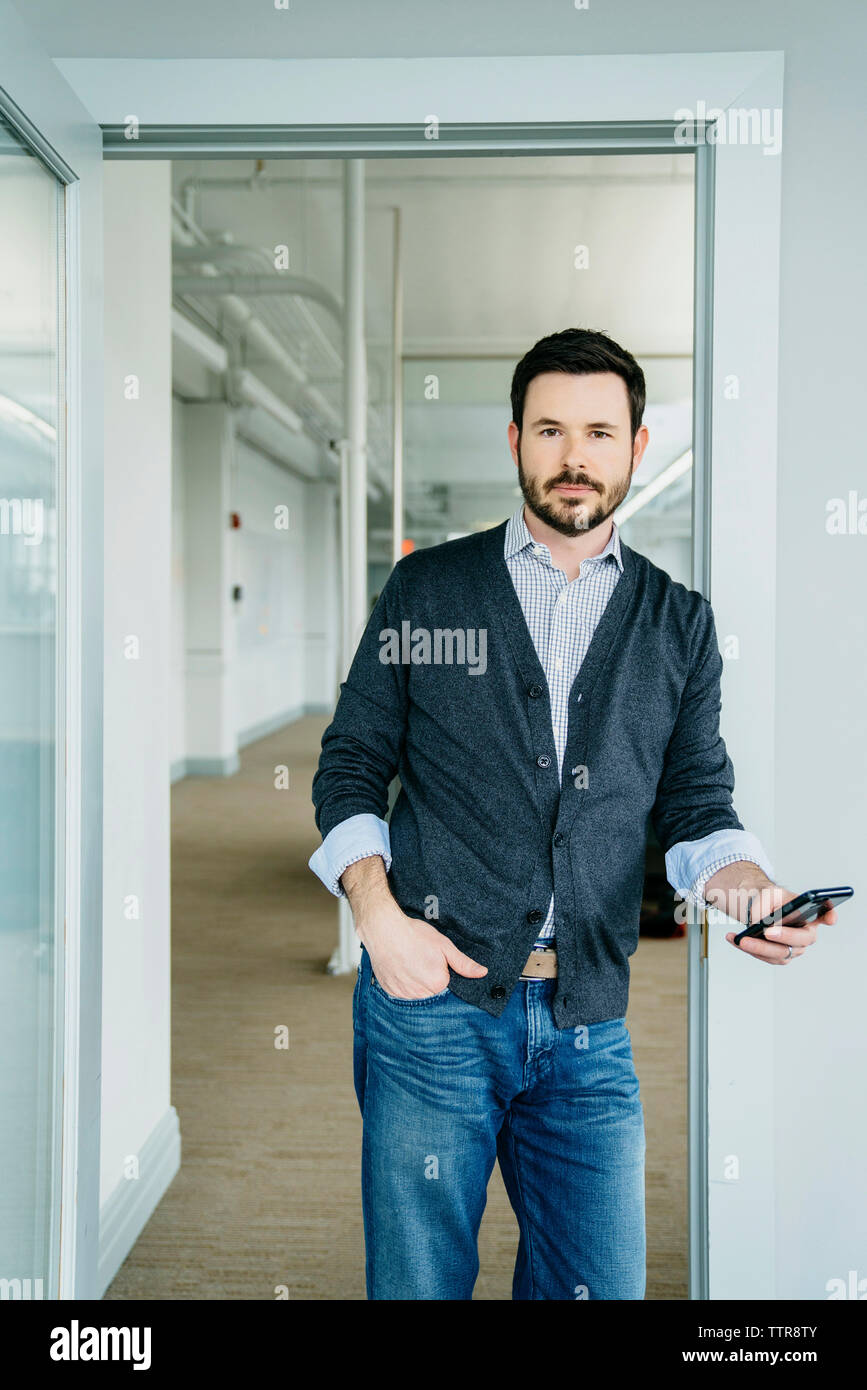 Portrait of businessman holding phone alors qu'il se tenait à l'embrasure in office Banque D'Images