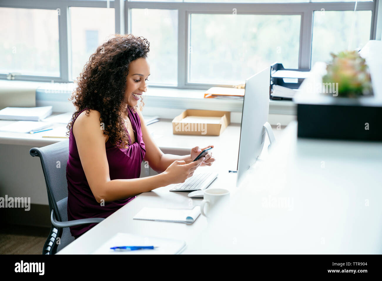 Smiling businesswoman using mobile phone while sitting at desk in office Banque D'Images