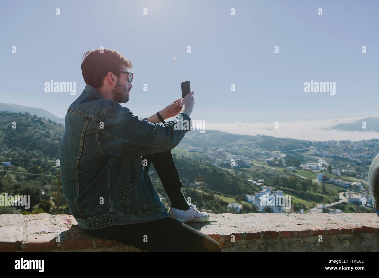 Side view of man photographing assis sur mur de retenue contre ciel clair au cours de journée ensoleillée Banque D'Images