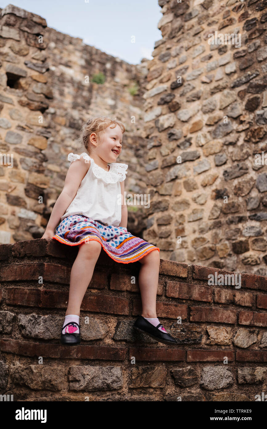 Low angle view of cute smiling girl aux cheveux blonds assis contre le mur en briques anciennes Banque D'Images