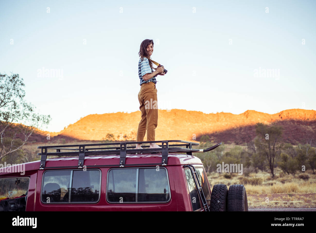 Portrait of smiling woman with camera debout sur toit de voiture contre un ciel clair Banque D'Images