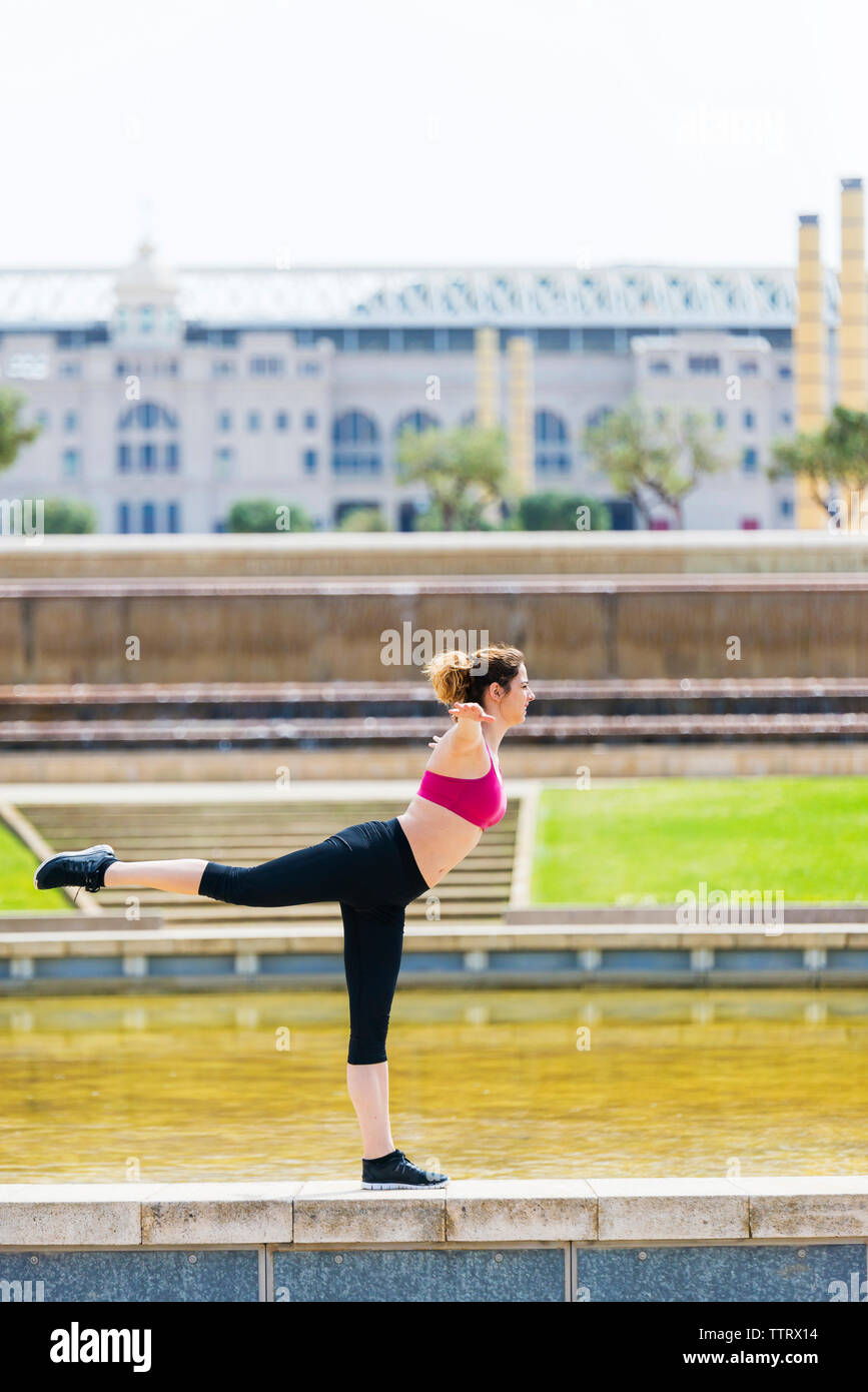 Vue latérale de l'athlète féminine avec bras tendus exerçant sur mur de retenue par pond at park Banque D'Images