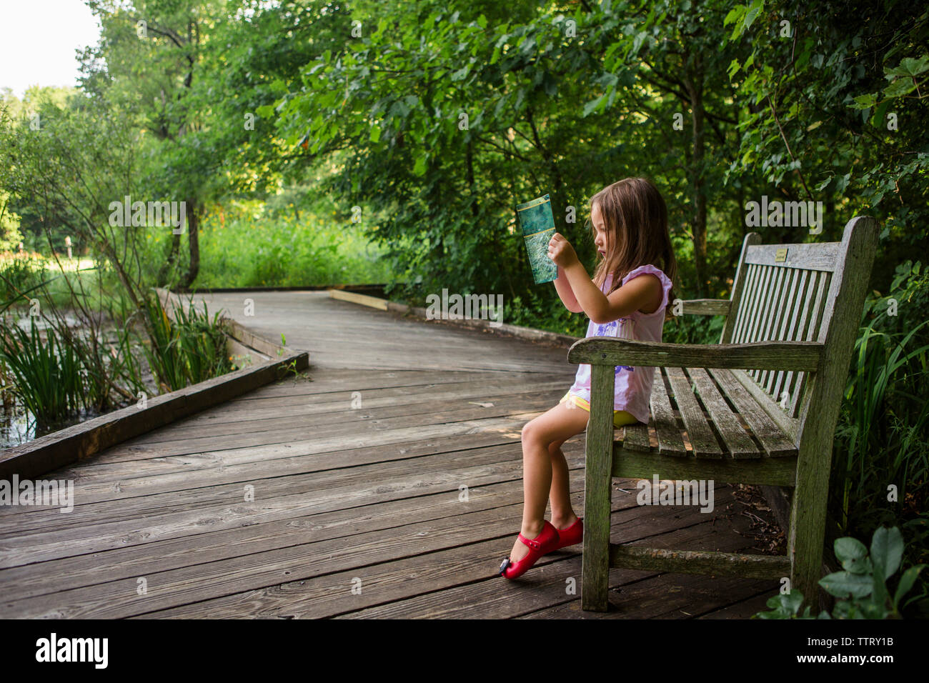 Vue latérale du girl reading book while sitting on bench at park Banque D'Images