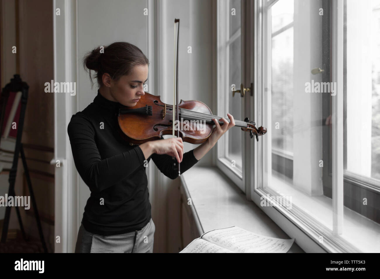 Femme confiante avec feuilles de musique sur appui de fenêtre à jouer du violon dans hôtel particulier Banque D'Images