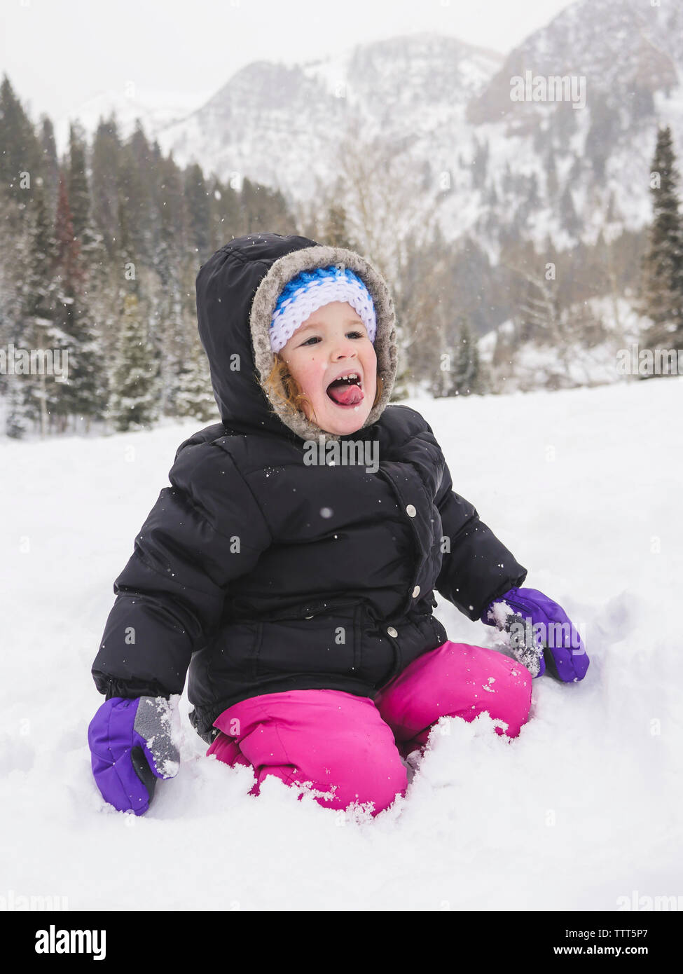 Happy baby girl à genoux sur le terrain couvert de neige contre mountain Banque D'Images