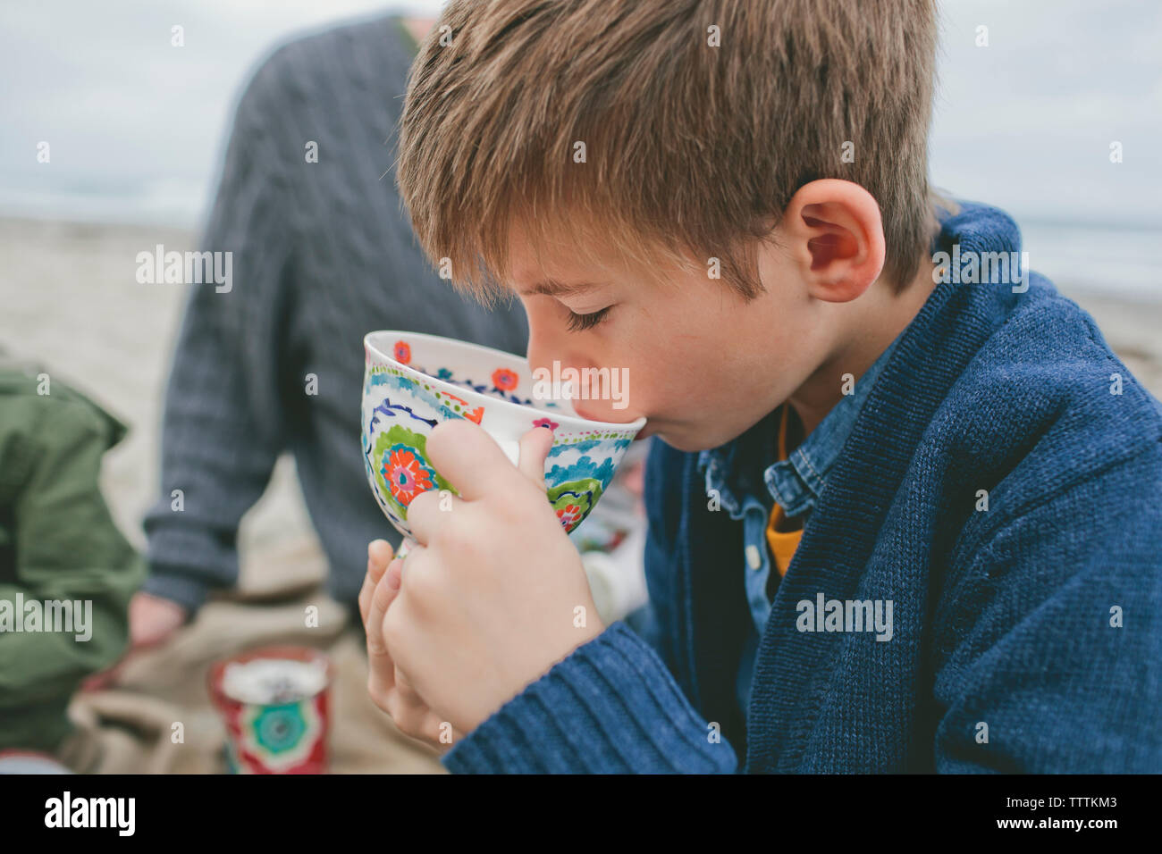 Boy drinking from cup à la plage avec la famille en arrière-plan Banque D'Images