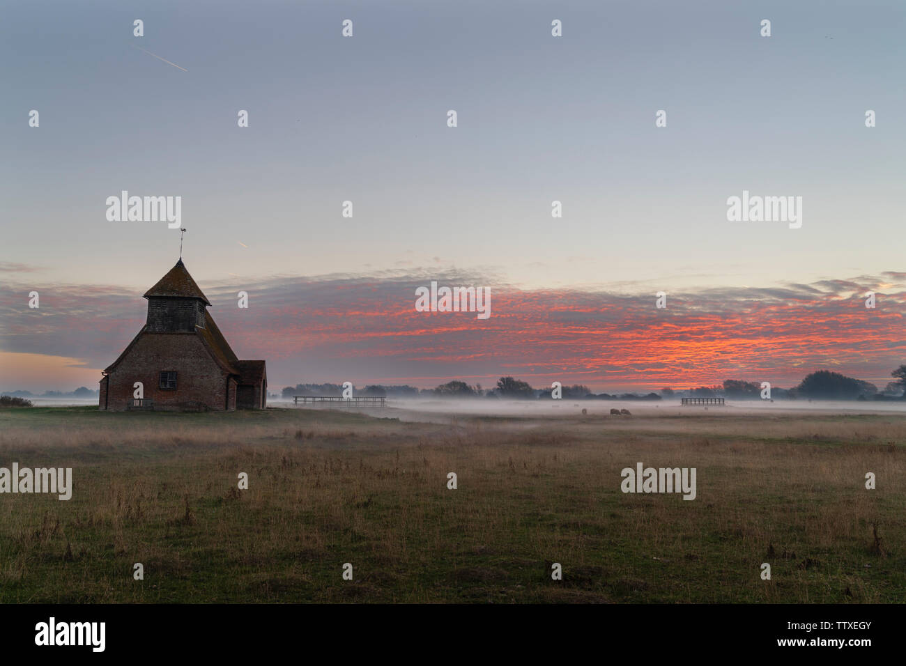 Saint Thomas Becket église à Romney Marsh. L'Église dans le marais à l'aube. Sol couvert de brouillard, nuages sur l'horizon prendre couleur dans la unrisen soleil. Banque D'Images