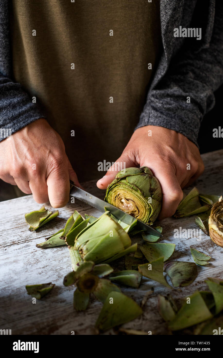 Closeup of a young man coupe avec un couteau de cuisine, des artichauts frais, sur une table en bois rustique blanc Banque D'Images