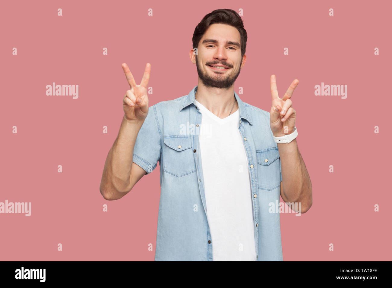 Portrait de jeune homme barbu heureux en bleu style casual shirt debout avec la victoire ou la paix part signer le geste et à la recherche à l'appareil photo avec smil dentelée Banque D'Images