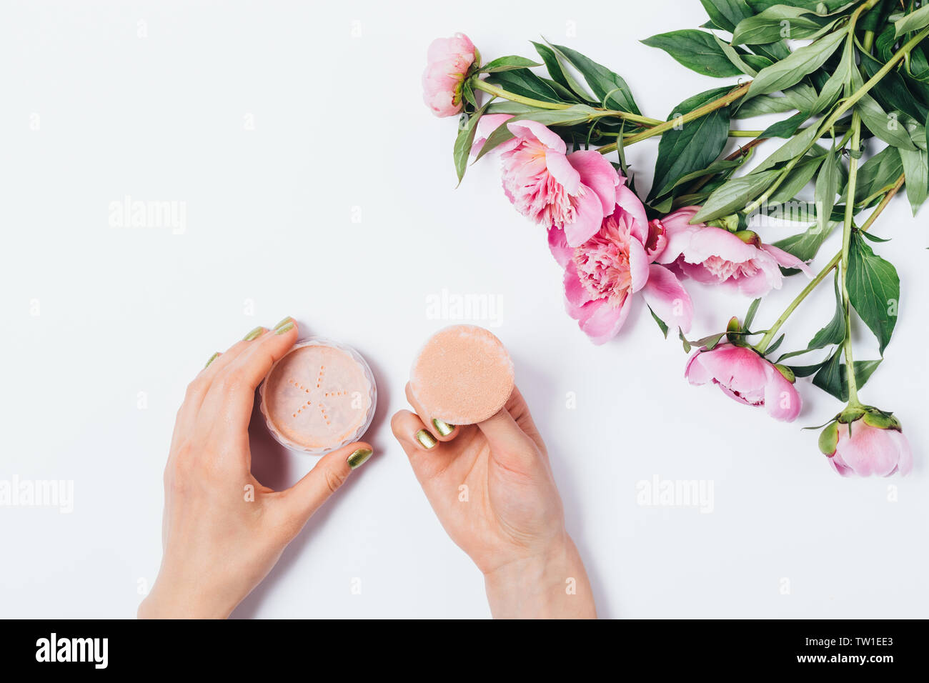 Top View woman's hands holding bouffée de maquillage avec applicateur poudre faciale beige lâche près de fresh bouquet de pivoines rose sur fond blanc, télévision lay. Banque D'Images