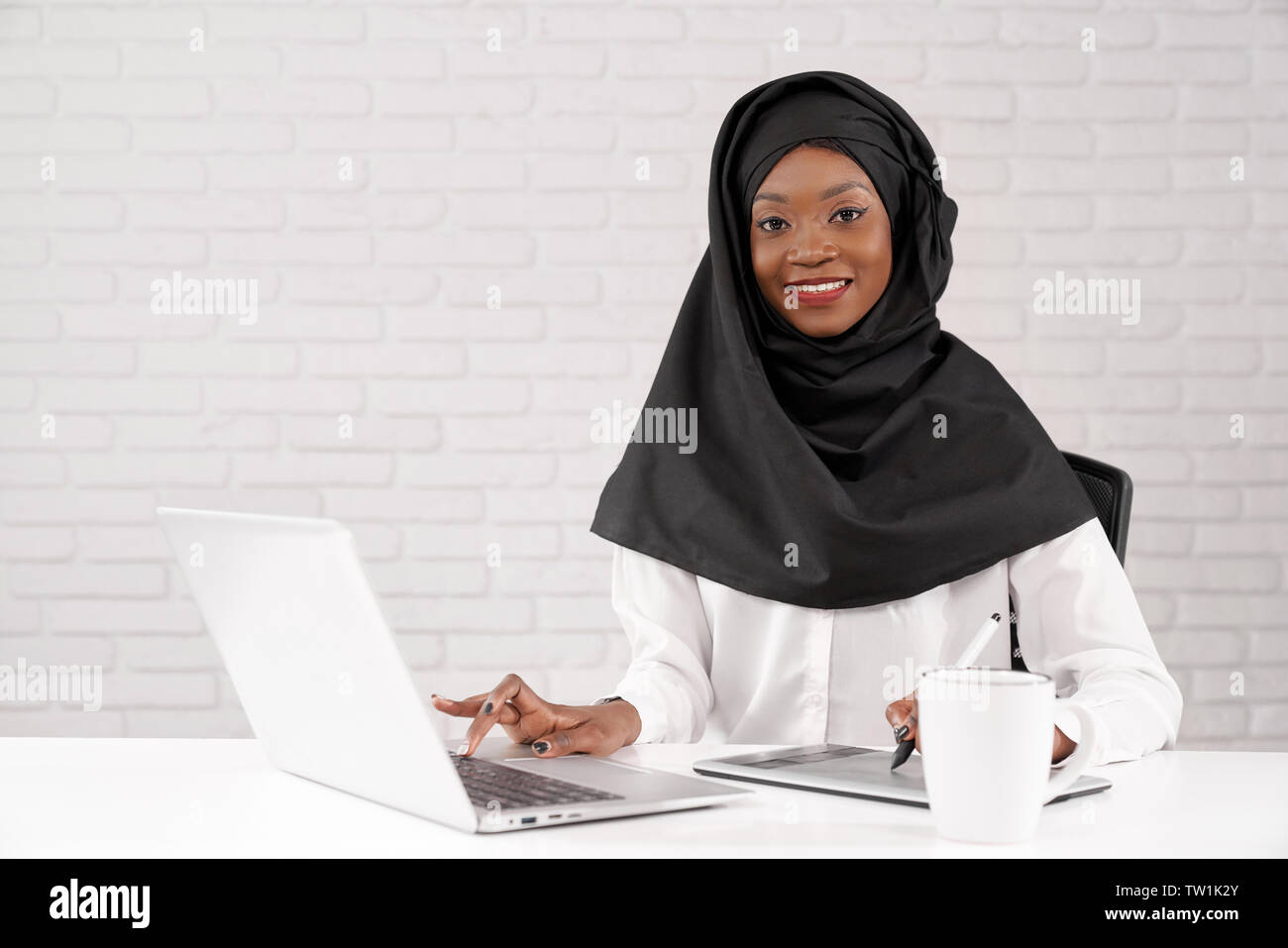 Les musulmans africains dame travaillant sur ordinateur dans office, looking at camera, souriant. Belle et jeune modèle africain assis à table blanche, à l'aide d'ordinateur portable. Banque D'Images