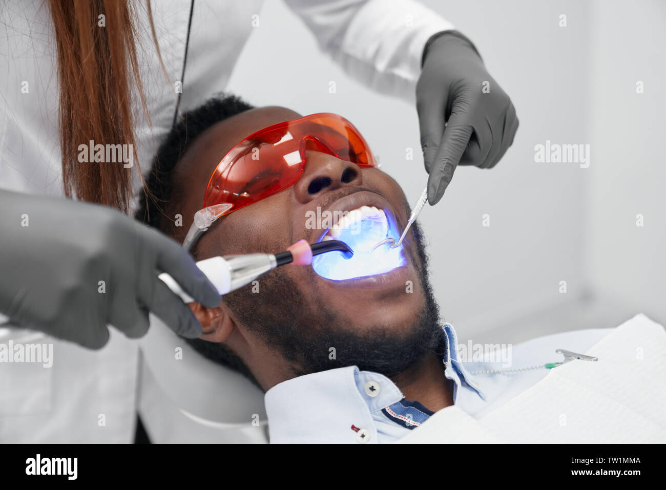 African man lying on dentiste président avec la bouche ouverte en lunettes de protection tandis que les femmes dentiste faisant remplissage avec de la lumière ultraviolette. Visite de l'homme médecin guérir les dents en clinique. Concept de stomatologie. Banque D'Images