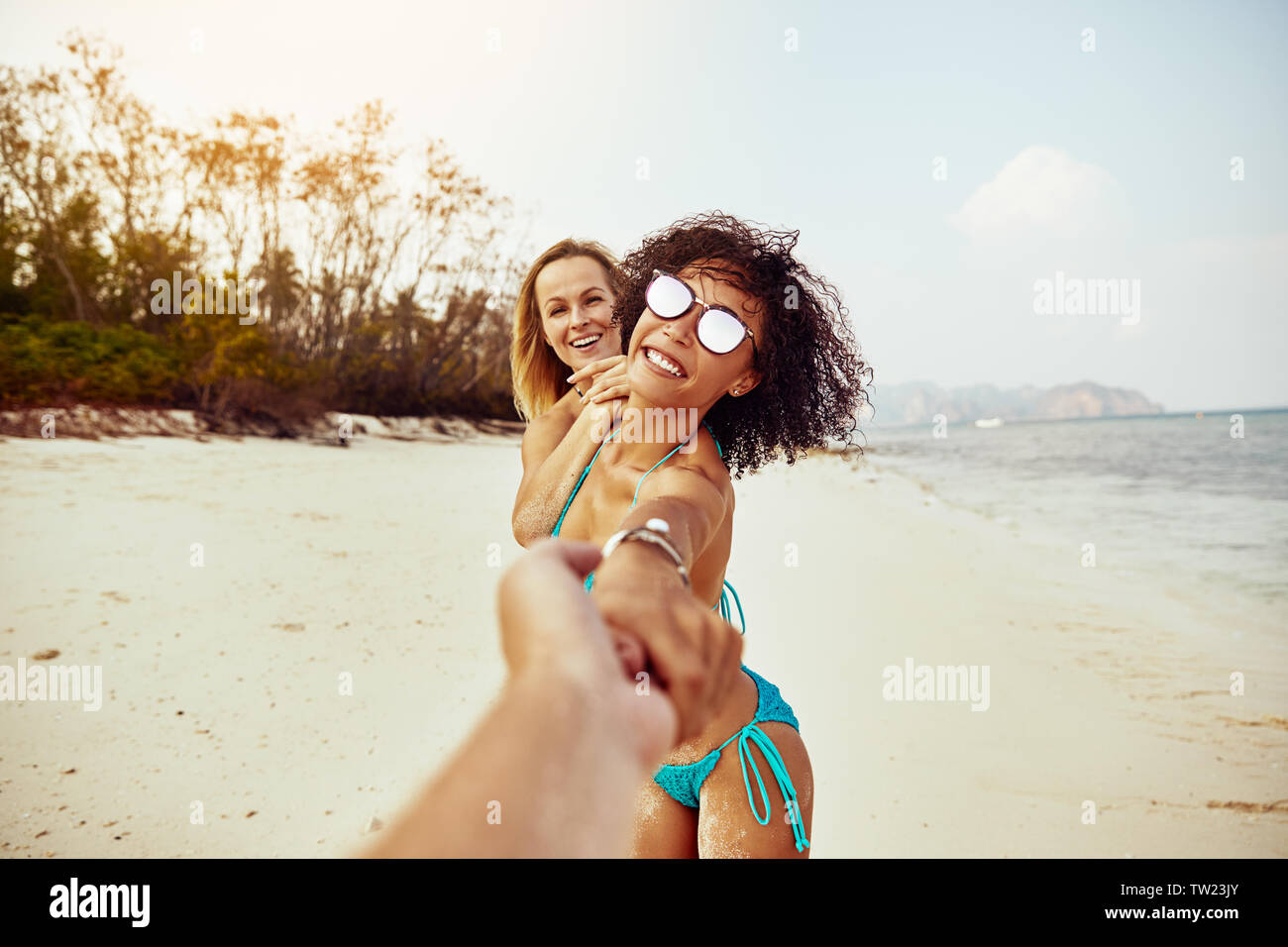 POV de rire deux jeunes femmes en bikini à la tête d'une personne par la main le long d'une plage de sable fin lors d'une vacances tropicales Banque D'Images