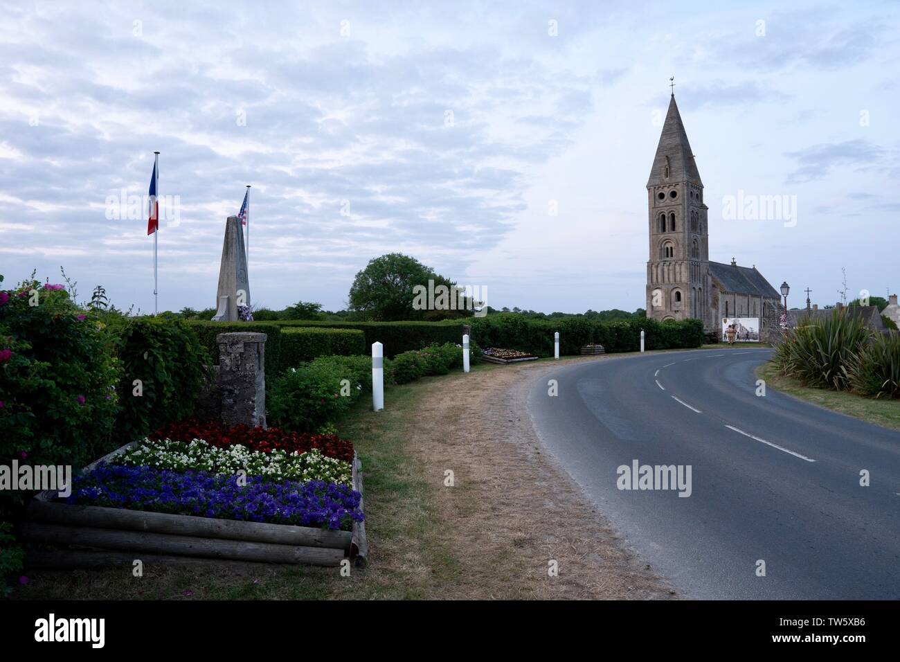 À l'entrée de l'église de Colleville sur Mer, un village français célèbre, vu depuis le côté de la route - WW2 memorial et fleurs en premier plan Banque D'Images