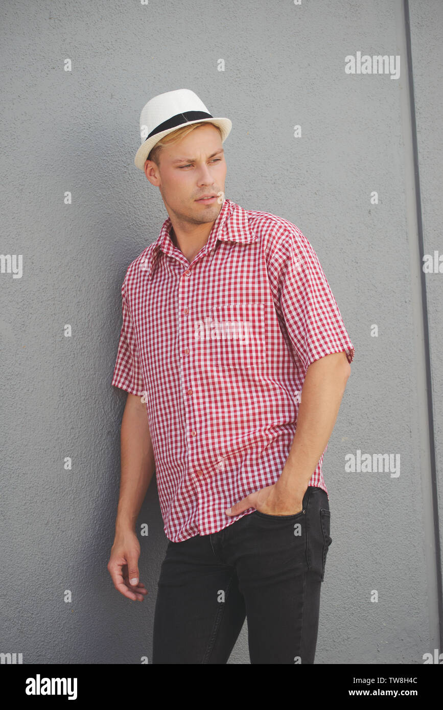 Un modèle masculin blond portant une chemise à carreaux rouge de l'Ouest, chapeau fedora blanc, posant contre un mur debout à l'extérieur, en regardant loin. Banque D'Images