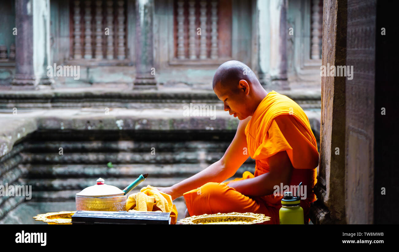 L'UNESCO, Angkor Wat, Siem Reap, Cambodge - Août 2016 : Une robe orange Un moine assis au bord du couloir et la préparation des aliments, avec un temple trouble b Banque D'Images