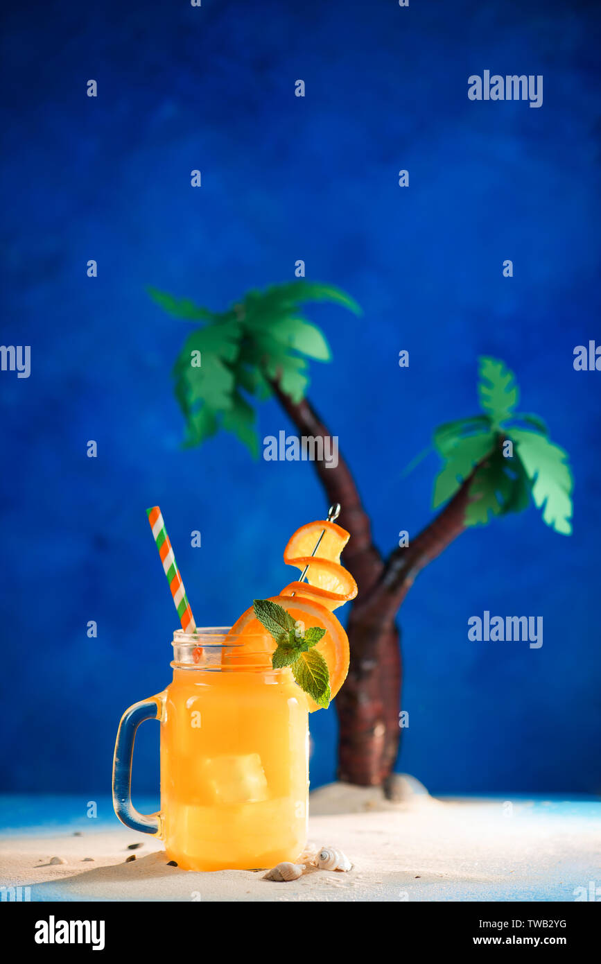 Cocktail tropical décorées dans un bocal en verre sur un fond bleu avec copie espace. Palmiers Papercraft avec jus jaune. Banque D'Images