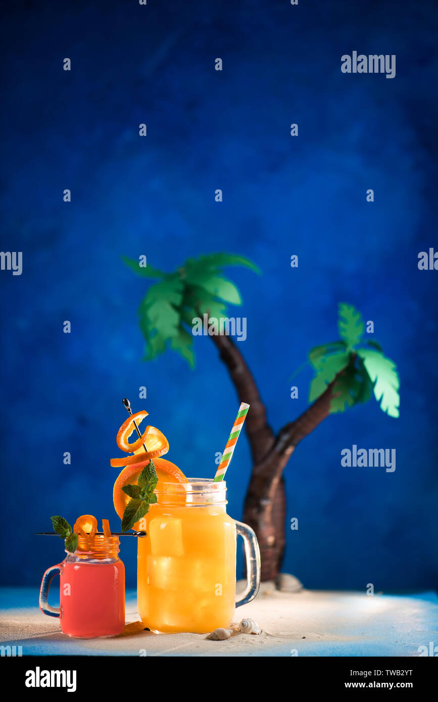 Des cocktails tropicaux décorées dans un bocal en verre sur un fond bleu avec copie espace. Palmiers Papercraft avec jus jaune et rouge. Banque D'Images