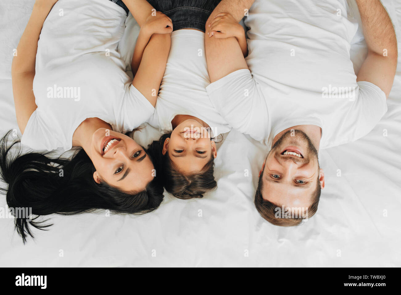 Famille heureuse allongé sur un lit blanc nouveau hugging et à la recherche à l'appareil photo, vue d'en haut. Mère, père et fille lying on bed at home Banque D'Images