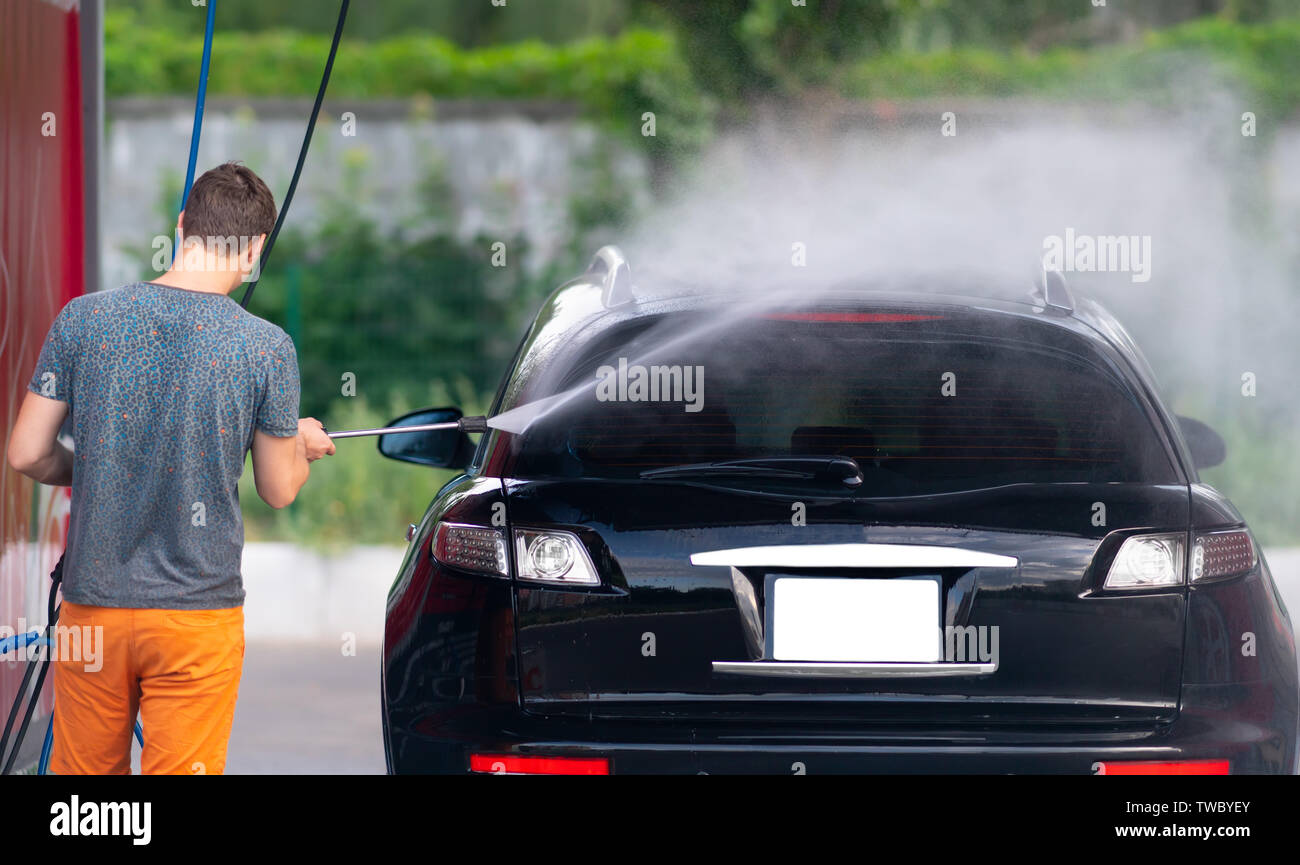 Jeune homme de pulvériser sa voiture avec de l'eau savonneuse pendant le nettoyage à un lave-auto à l'aide d'une buse haute pression Banque D'Images