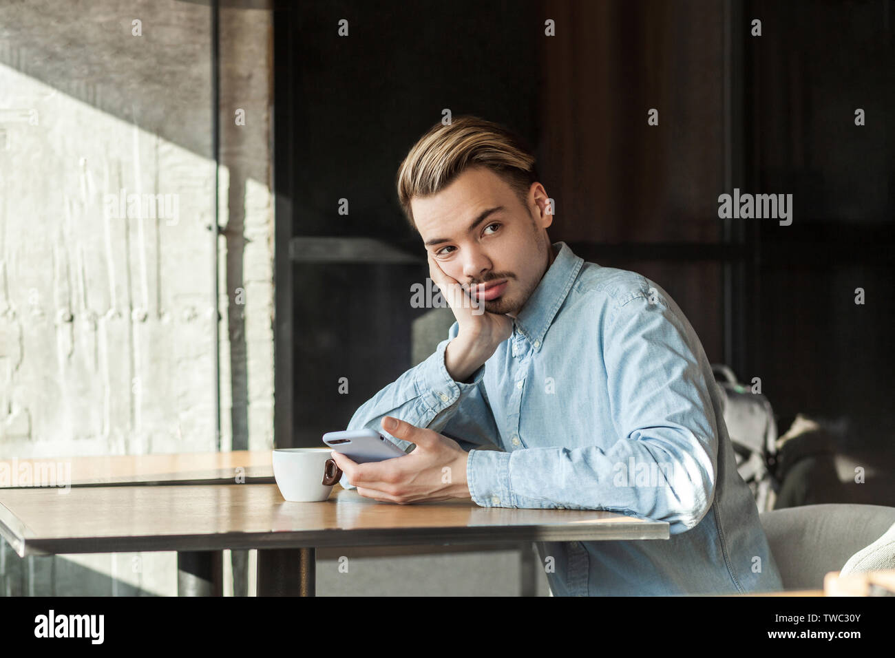 Portrait de beau jeune homme barbu réfléchie dans la chemise en denim bleu assis dans un café et de boire du café, fatigué de l'attente de tes amis de respect ou de thi Banque D'Images
