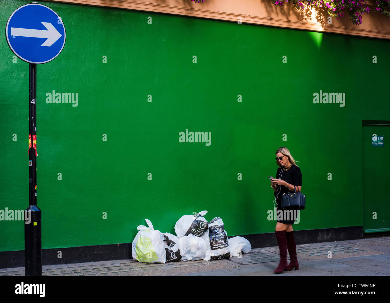 Jeune femme exerçant son sac à main et l'utilisation de smartphone pendant la marche verte passé mur peint avec des sacs poubelle en face de lui dans le centre de Londres. Banque D'Images