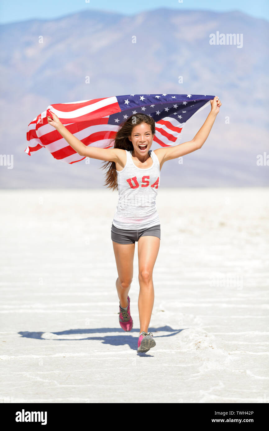 Femme de l'athlète avec le drapeau américain en marche avec USA T-shirt montrant large geste excité et heureux à l'extérieur de la nature du désert. Cheerful woman cheering gagnant de remise en forme. Banque D'Images