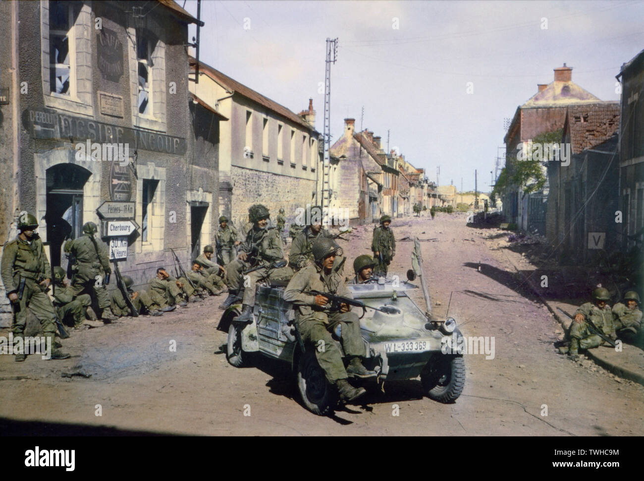 Parachutistes américains en jeep allemande patrouillant Carentan tandis que d'autres soldats américains reste contre un bâtiment, Normandie, France, juin 1944 Banque D'Images