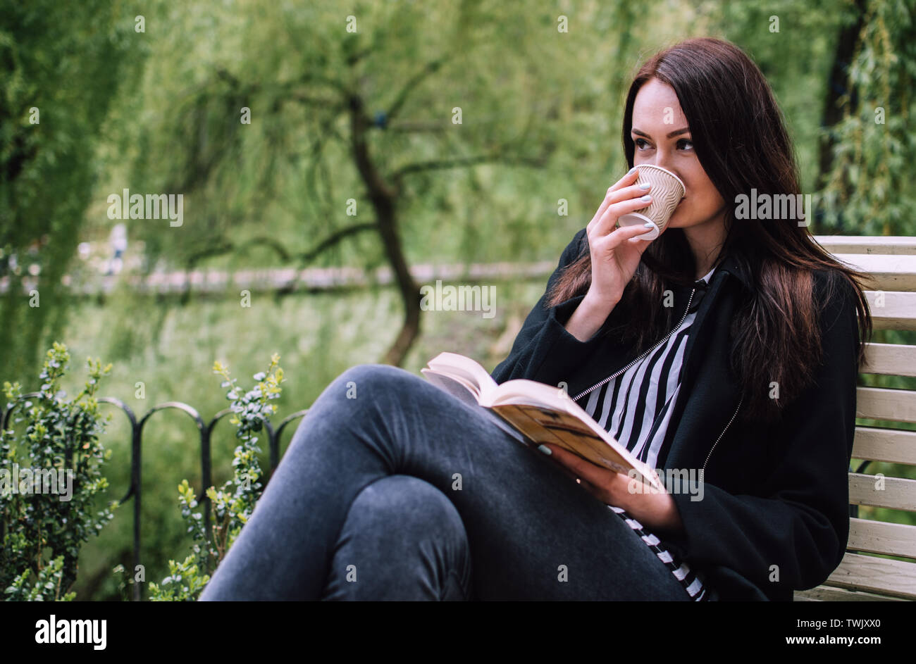 Smiling brunette fille assise sur un banc dans un parc lorsque boire du café dans une tasse de papier et regardant au loin. Livre à couverture souple dans ses mains. Banque D'Images
