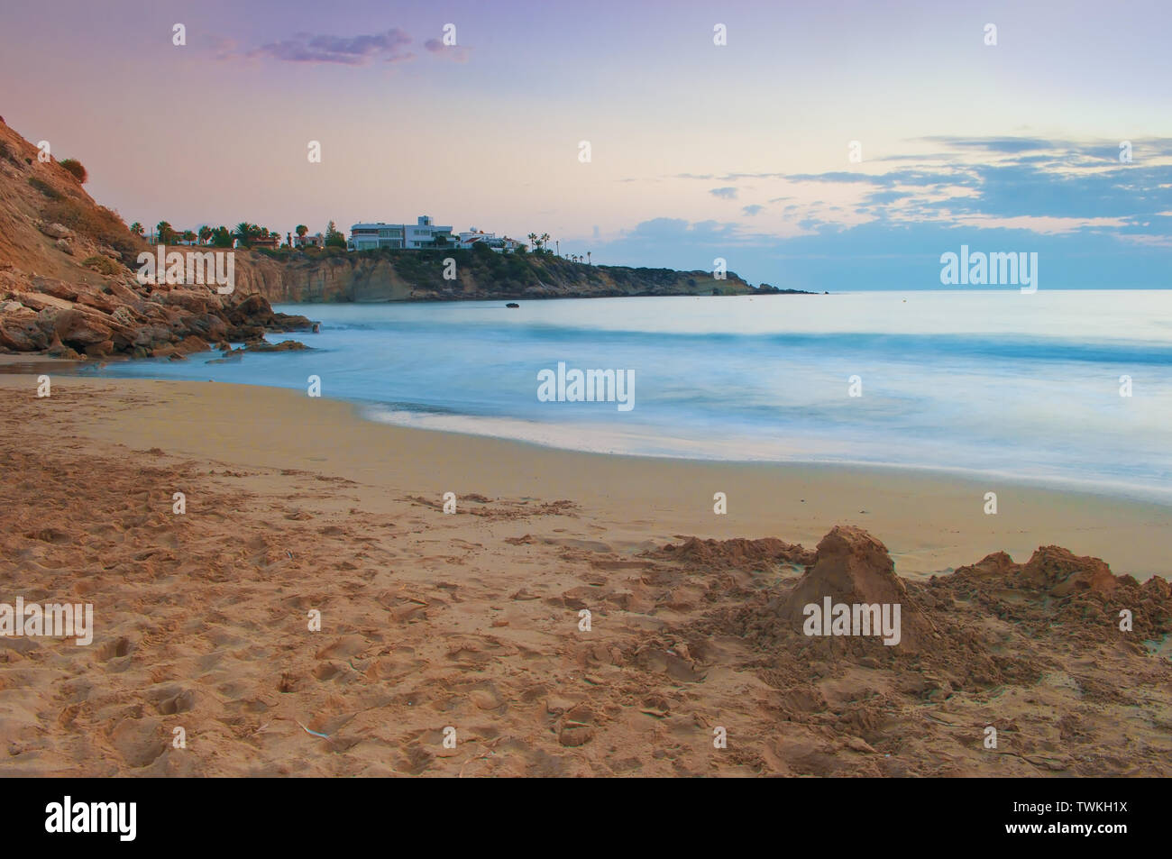 Vue de la plage de sable de la baie de Corail vide près de Paphos, Chypre. Le coucher du soleil, ciel rose bleu clair au-dessus de l'eau peu profonde avec des vagues, des villas sur la falaise. Chaude soirée Banque D'Images