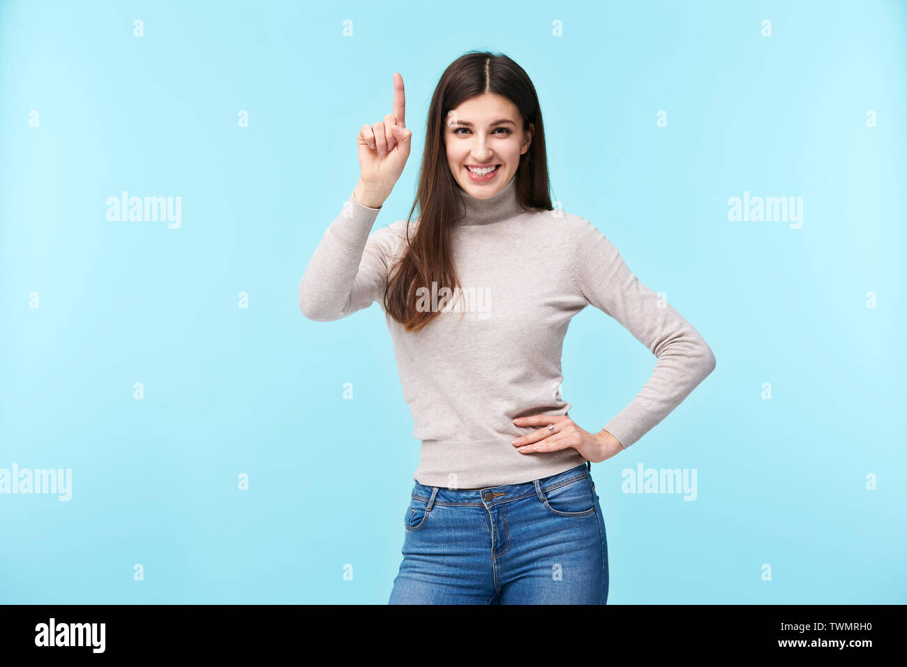 Studio portrait d'une jolie femme caucasienne, doigt pointant vers le haut, smiling at camera, bras akimbo, isolé sur fond bleu Banque D'Images