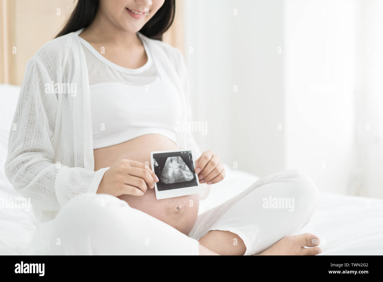 Femme enceinte tenant la photo de l'échographie sur son ventre dans le lit à la Chambre. Arrière-plan blanc. Des soins de santé prénatale concept. Banque D'Images