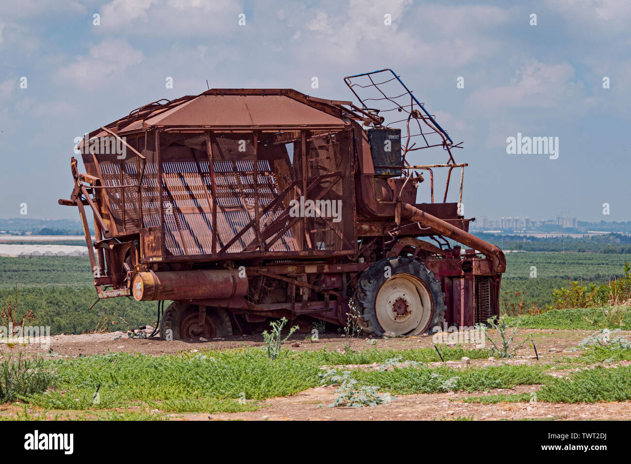 A broken down rusty old machine cueillette du coton se trouve au-dessus de champs et de serres modernes en Israël avec une ville palestinienne dans l'arrière-plan Banque D'Images