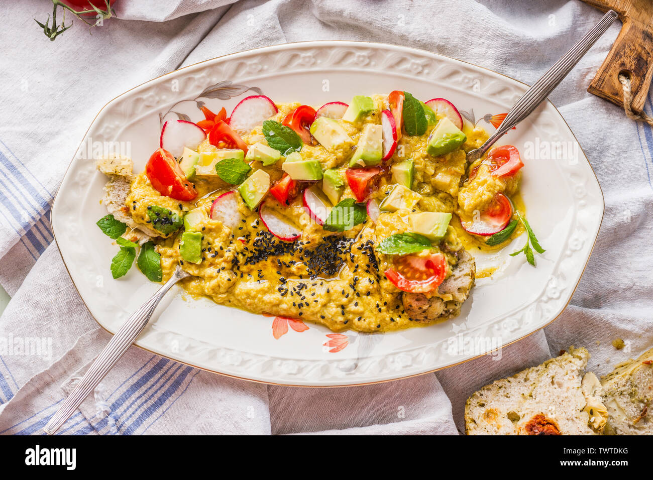 La plaque de l'hummus garni d'huile d'olive, avocat, légumes et herbes fraîches hachées sur table de cuisine avec fourchette et pain, vue d'en haut. La nourriture végane concept. Il Banque D'Images