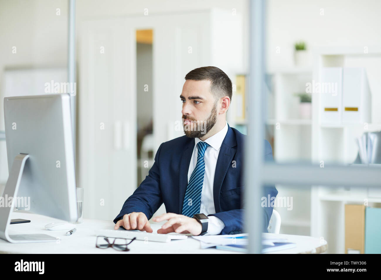 Young businessman sérieux barbu en costume bleu foncé assis à table et la saisie sur clavier d'ordinateur tout en analysant des données statistiques Banque D'Images