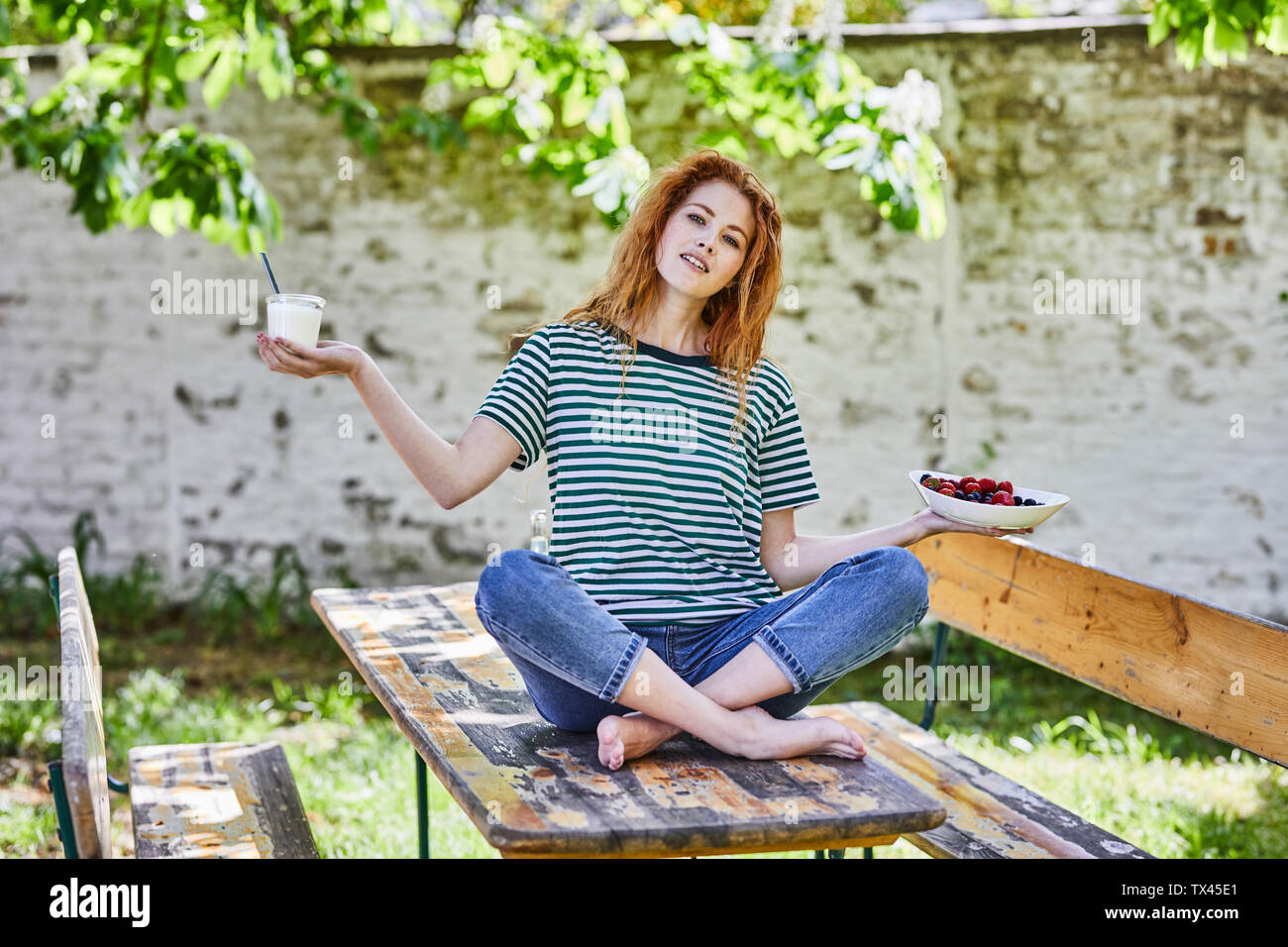 Portrait of young woman sitting on wooden table in garden holding bol de fruits et un verre de yaourt Banque D'Images