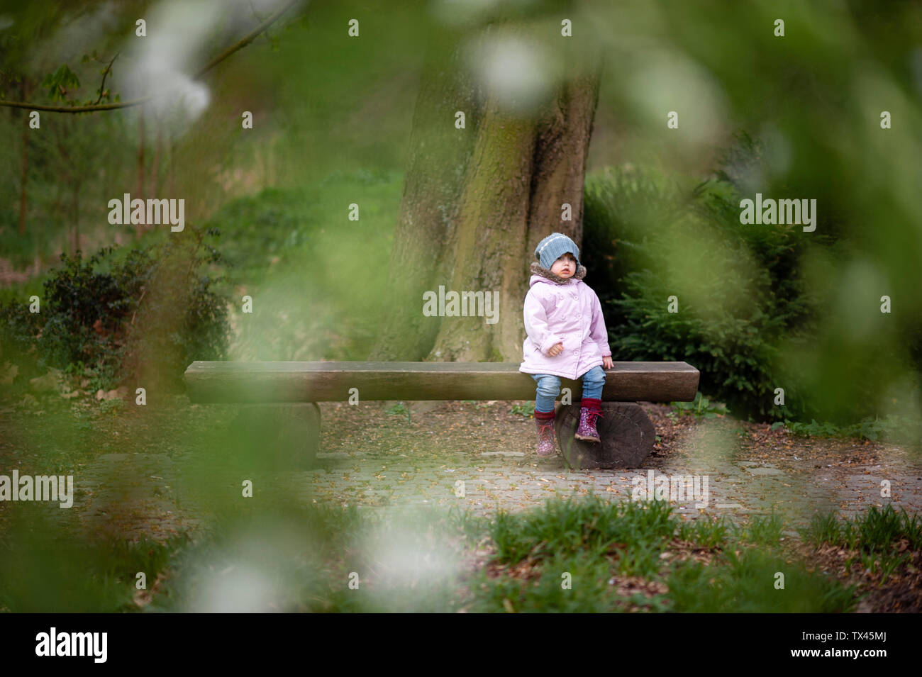 Little girl wearing blue hat manteau rose et assis sur un banc dans un parc Banque D'Images